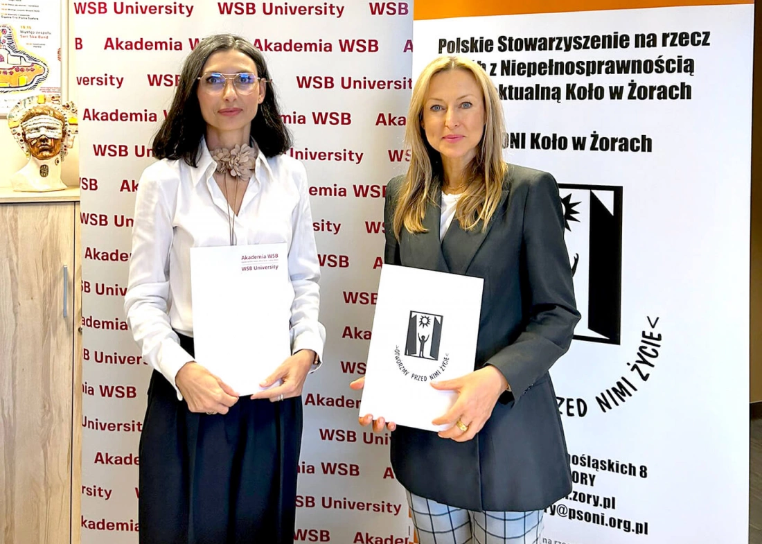 Dwie kobiety trzymają dokumenty, stojąc przed bannerami Akademii WSB i Polskiego Stowarzyszenia na rzecz osób z Niepełnosprawnością.