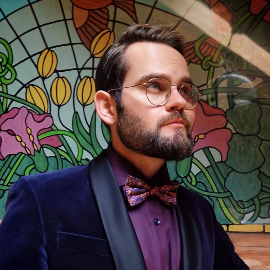 Mężczyzna w eleganckim garniturze z muszką, stojący przed kolorowym witrażem.