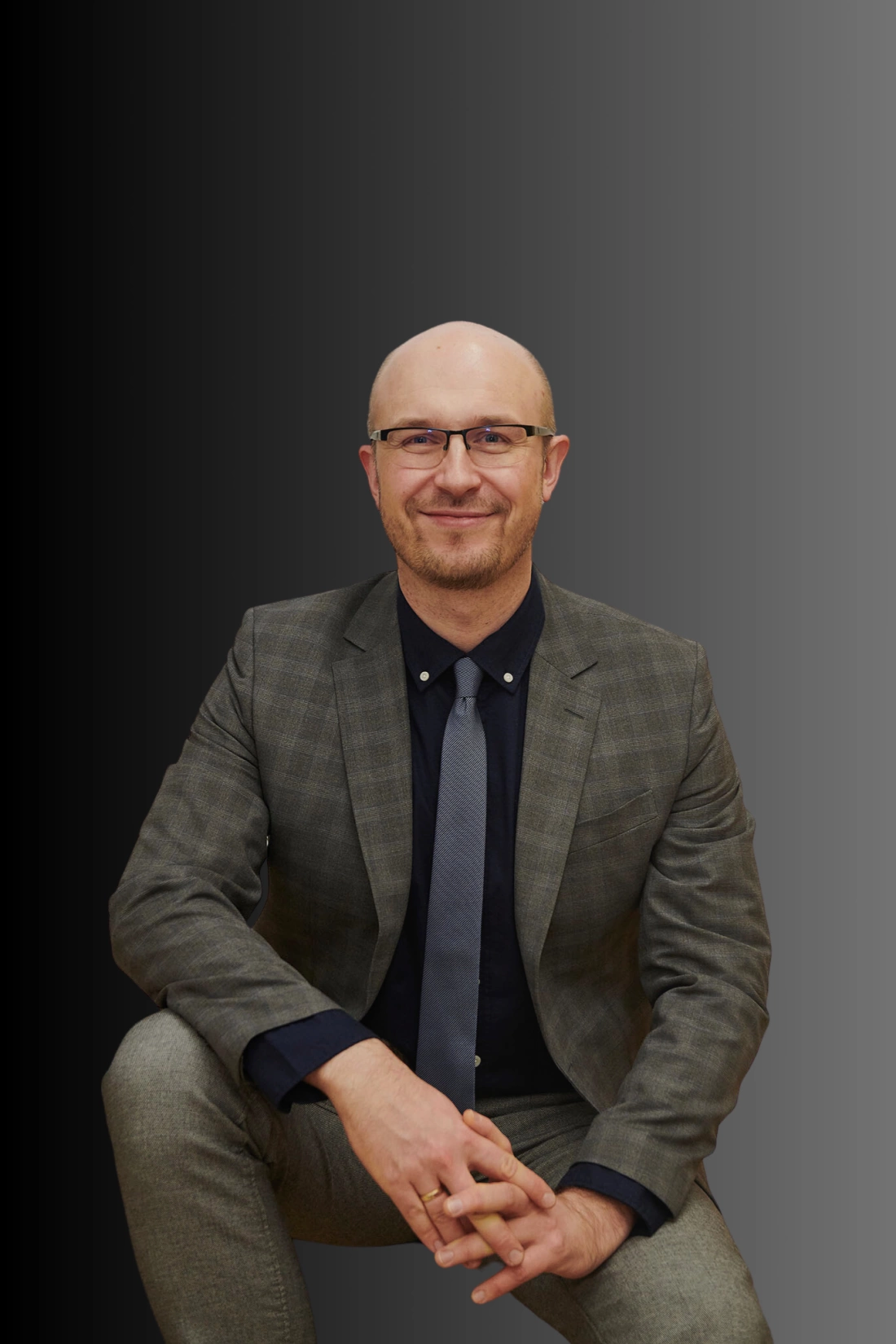 Mężczyzna w eleganckim garniturze, siedzący na jednym kolanie, z uśmiechem na twarzy.