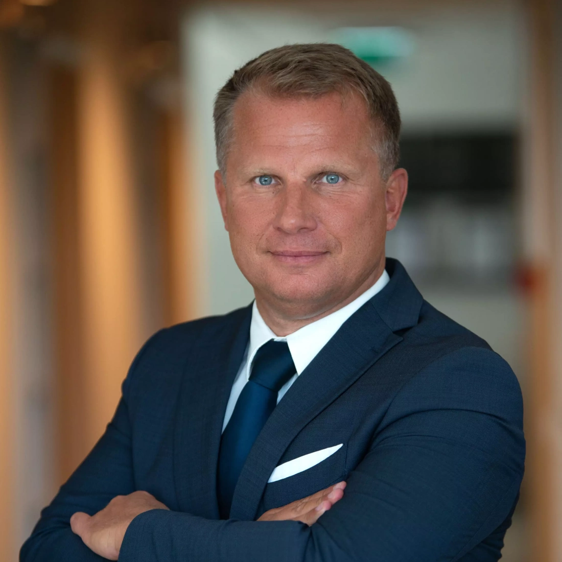 Mężczyzna w garniturze z niebieskim krawatem, z założonymi rękami, uśmiecha się do kamery. Tło biurowe.