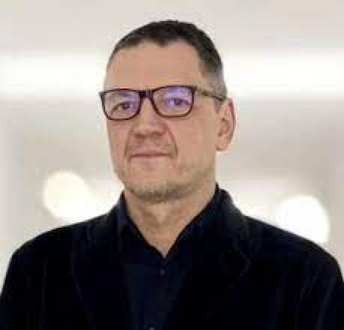 Mężczyzna w okularach, w jasnym tle, ubrany w czarną marynarkę.