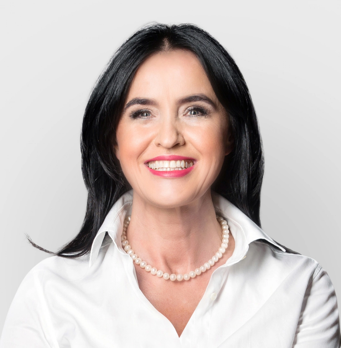 Kobieta z długimi, ciemnymi włosami, uśmiechająca się w białej koszuli i perłowej kolii.