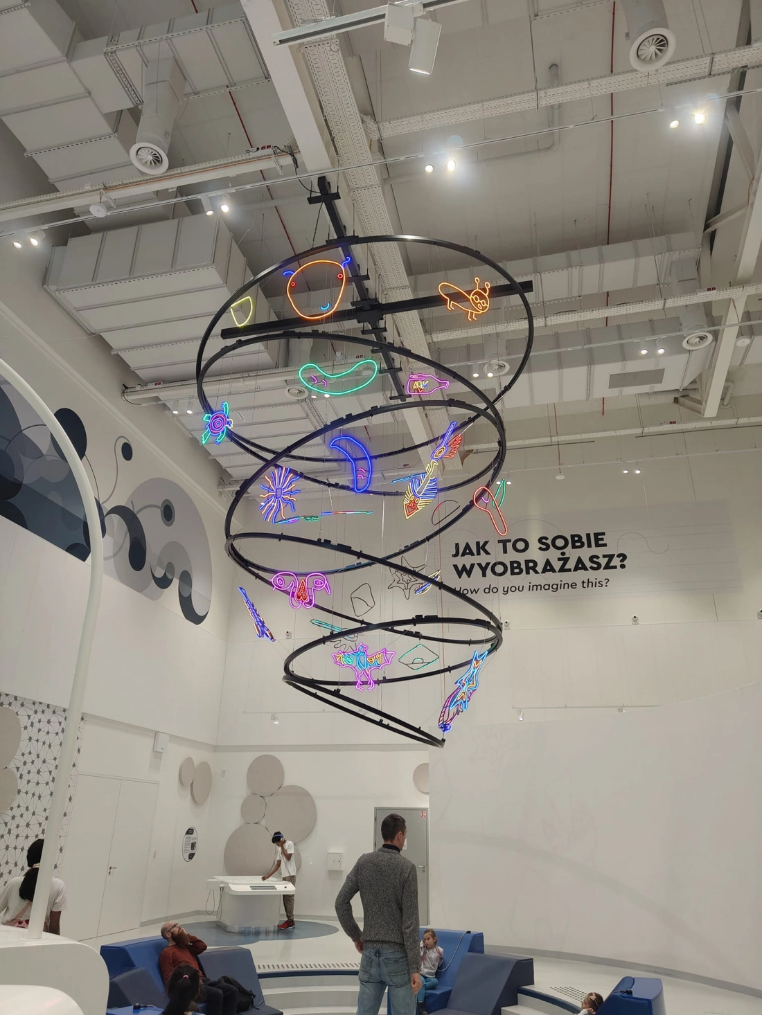 Nowoczesna przestrzeń z wiszącą instalacją świetlną w kształcie zwierząt, a w tle tekst "Jak to sobie wyobrażasz?"