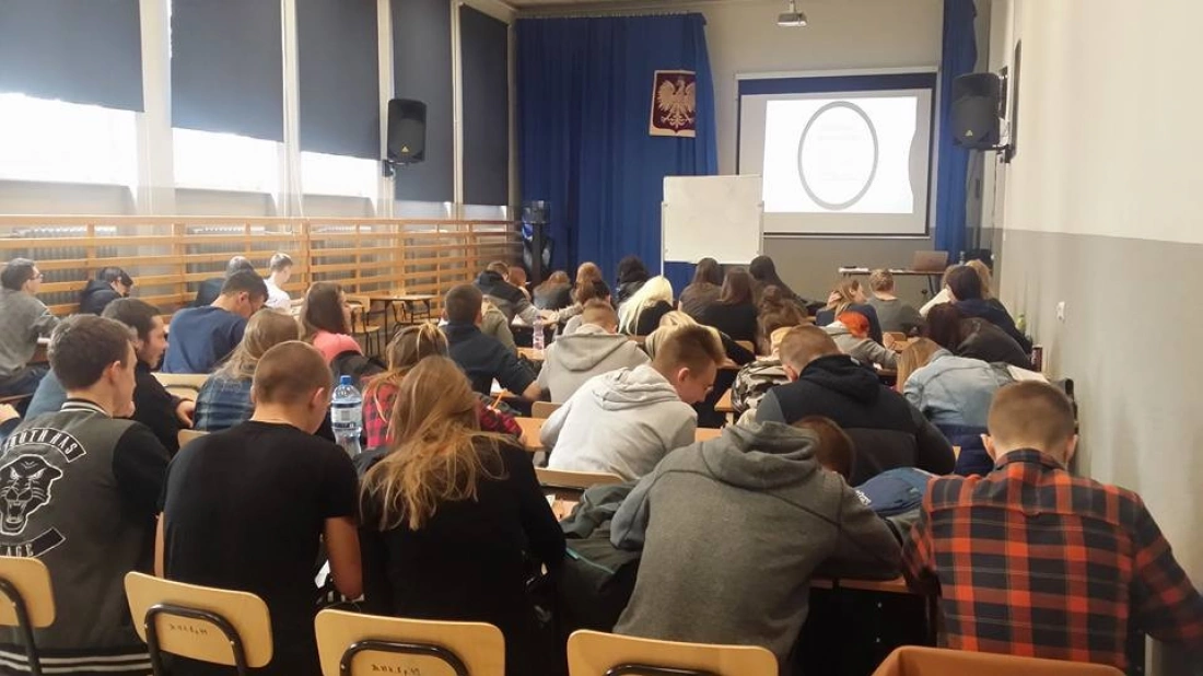 Sala lekcyjna z uczniami siedzącymi przy stolikach, wpatrzonymi w ekran prezentacji.