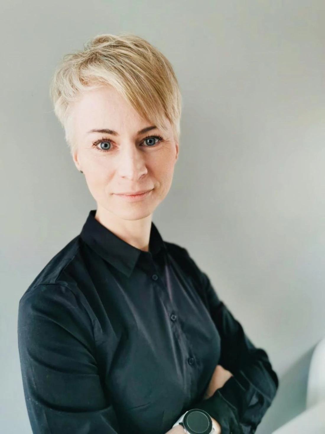 Kobieta o krótkich, jasnych włosach, ubrana w czarną koszulę, stoi z założonymi rękami na tle szarej ściany.