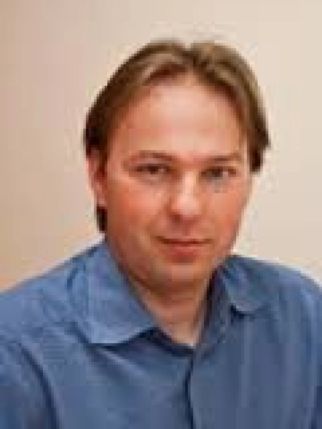 Mężczyzna z brązowymi włosami, ubrany w niebieską koszulę, na tle jasnej ściany.