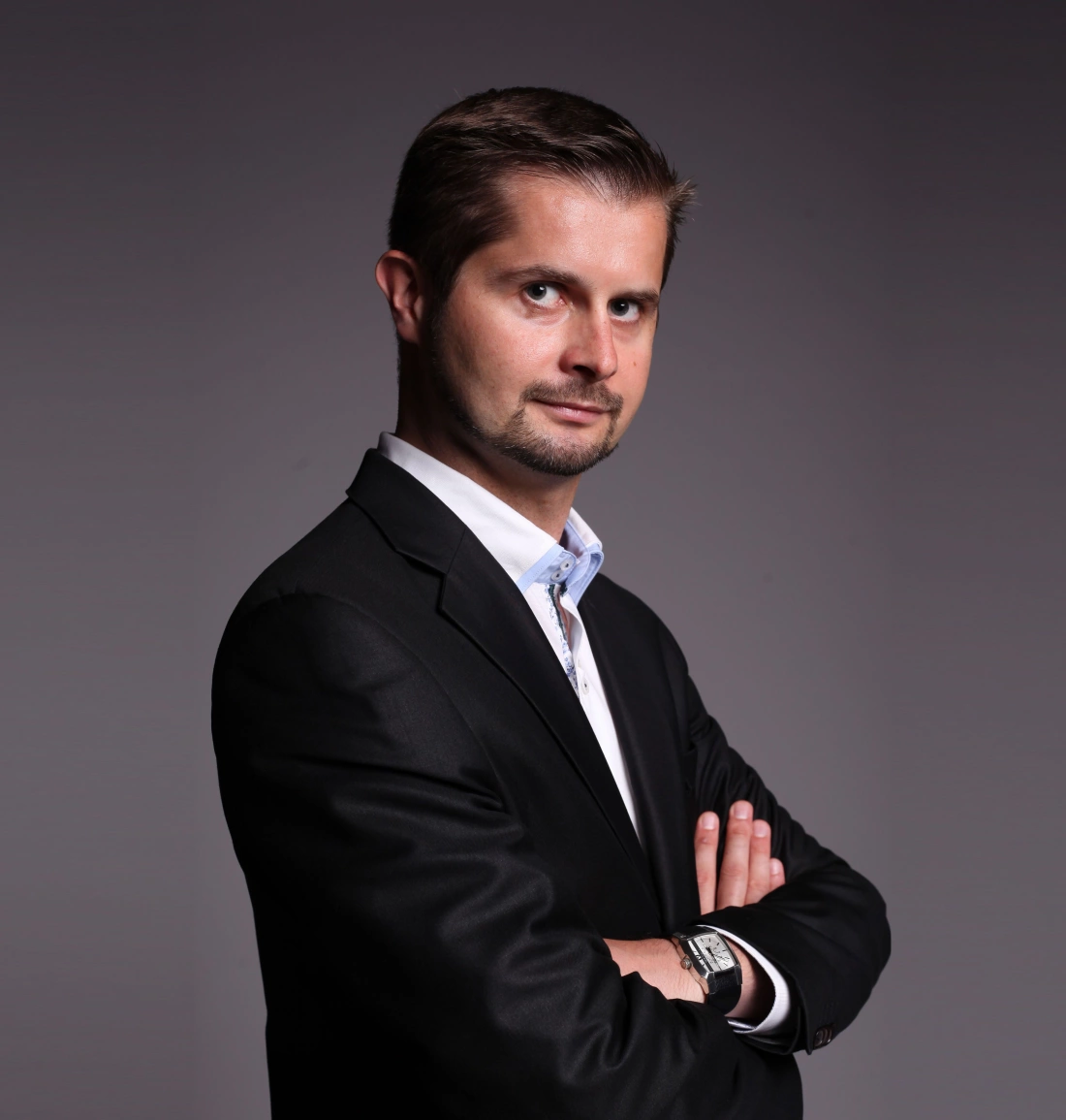 Mężczyzna w eleganckim garniturze, z założonymi rękami, patrzy w obiektyw.