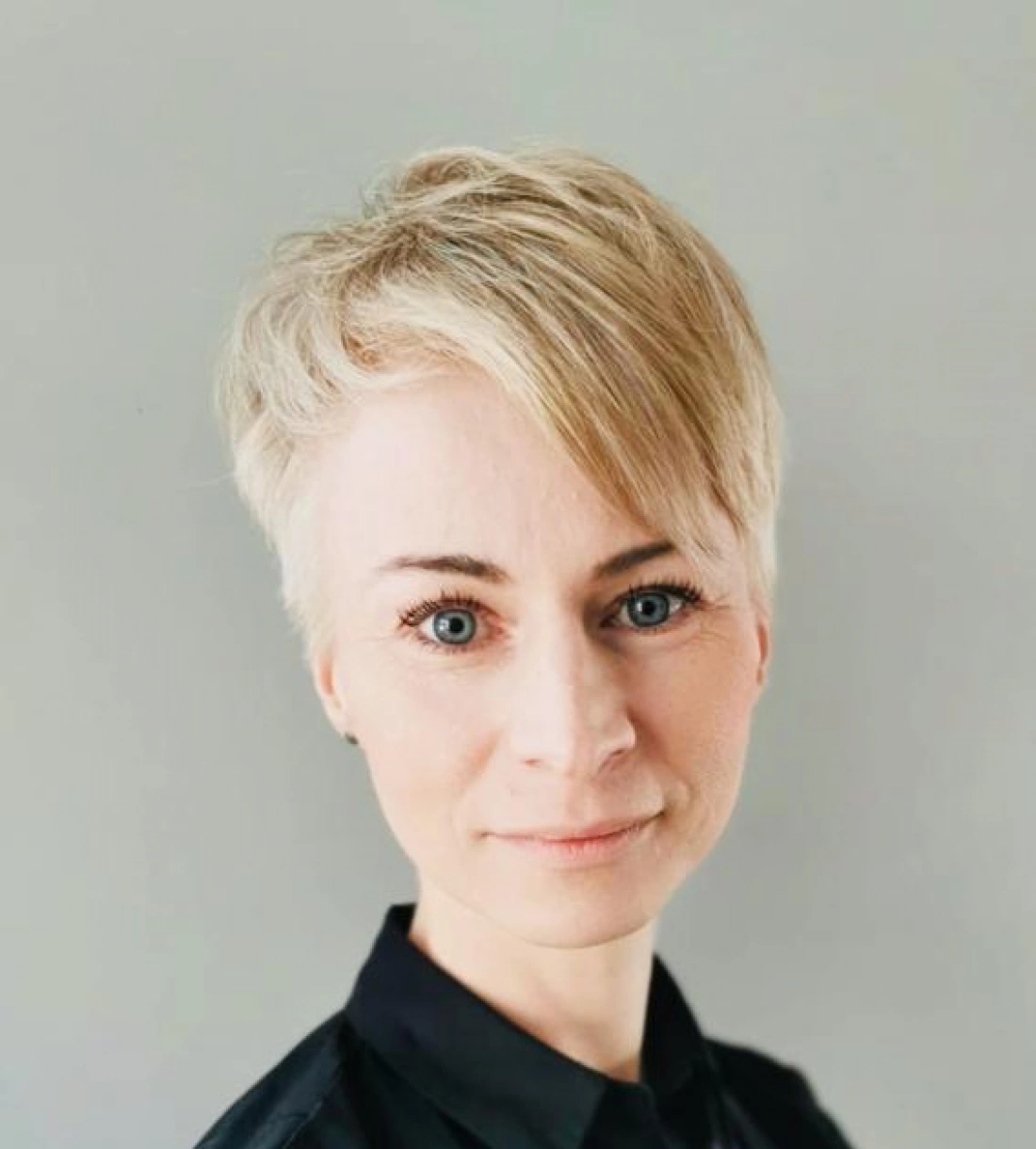 Kobieta z krótkimi, jasnymi włosami i niebieskimi oczami, uśmiechająca się lekko na tle szarej ściany.