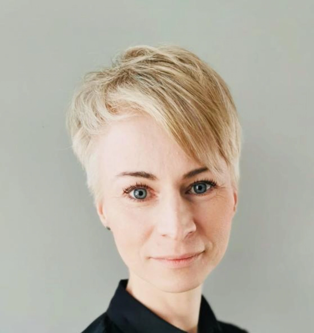 Kobieta z krótkimi, jasnymi włosami i niebieskimi oczami, ubrana w czarną bluzkę, na tle szarej ściany.