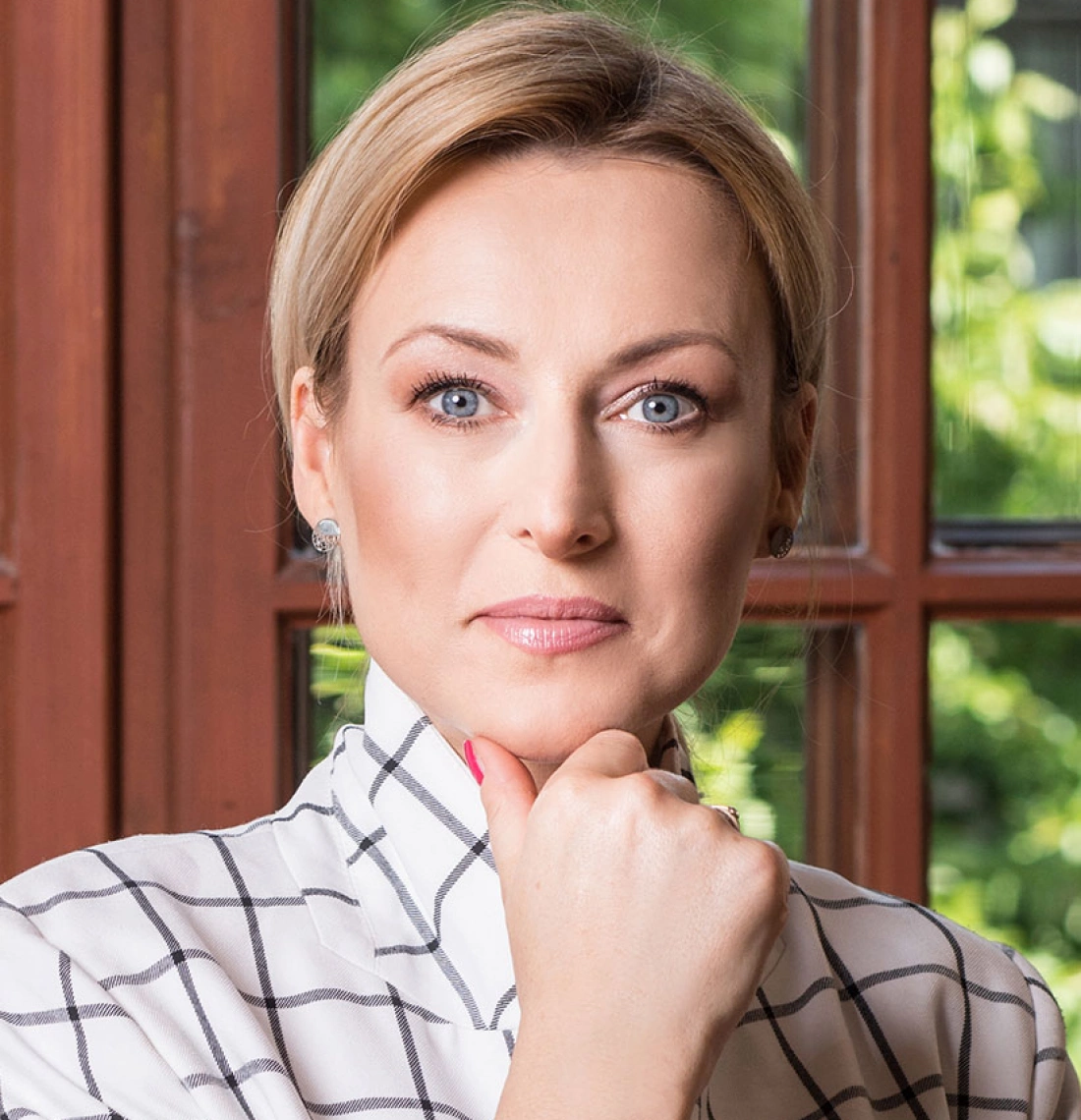 Kobieta o jasnych włosach i niebieskich oczach, ubrana w koszulę w kratę, z ręką przy brodzie, stoi przy oknie.
