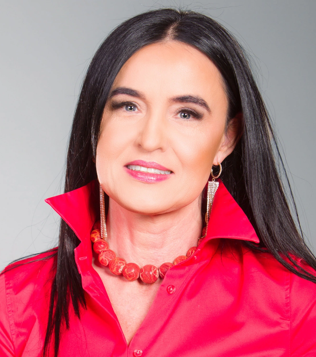 Kobieta z długimi, ciemnymi włosami, uśmiechająca się w czerwonej koszuli z podniesionymi kołnierzykami, ozdobiona naszyjnikiem i dużymi kolczykami.