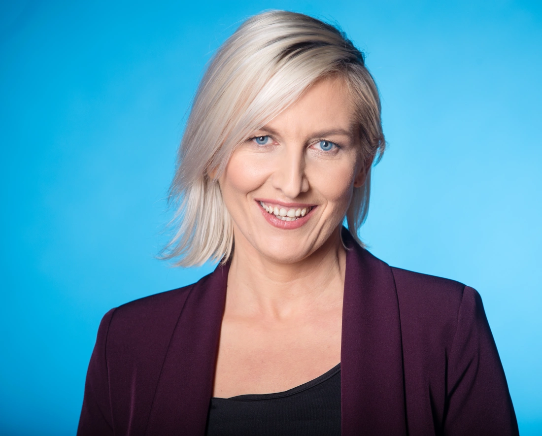 Portret kobiety z krótkimi blond włosami, uśmiechającej się na tle niebieskiego tła. Ubrana w ciemną bluzkę i żakiet.