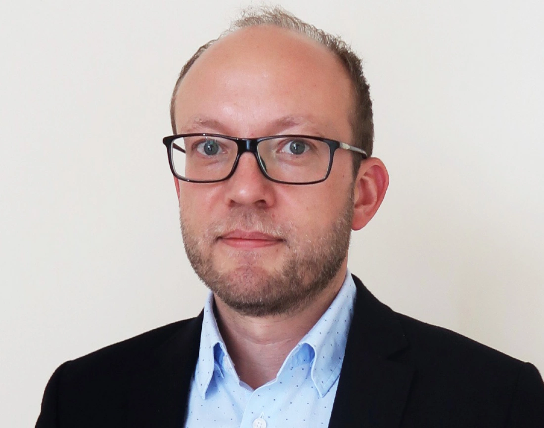 Mężczyzna w okularach, w ciemnej marynarce, na jasnym tle.