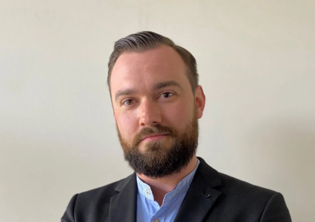 Mężczyzna w garniturze z zarostem, stojący na tle jednolitej ściany.