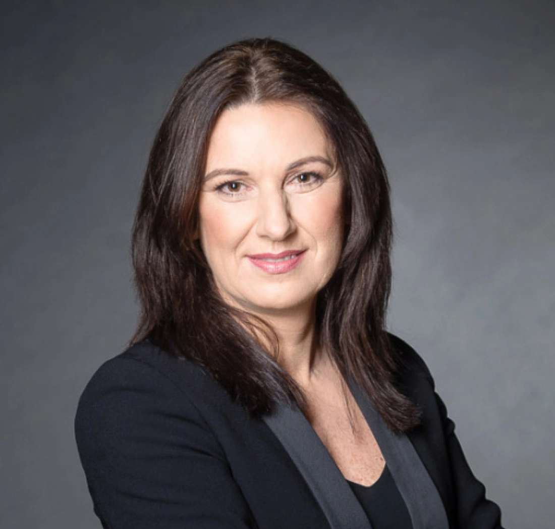 Portret uśmiechniętej kobiety w czarnej marynarce na tle szarym.