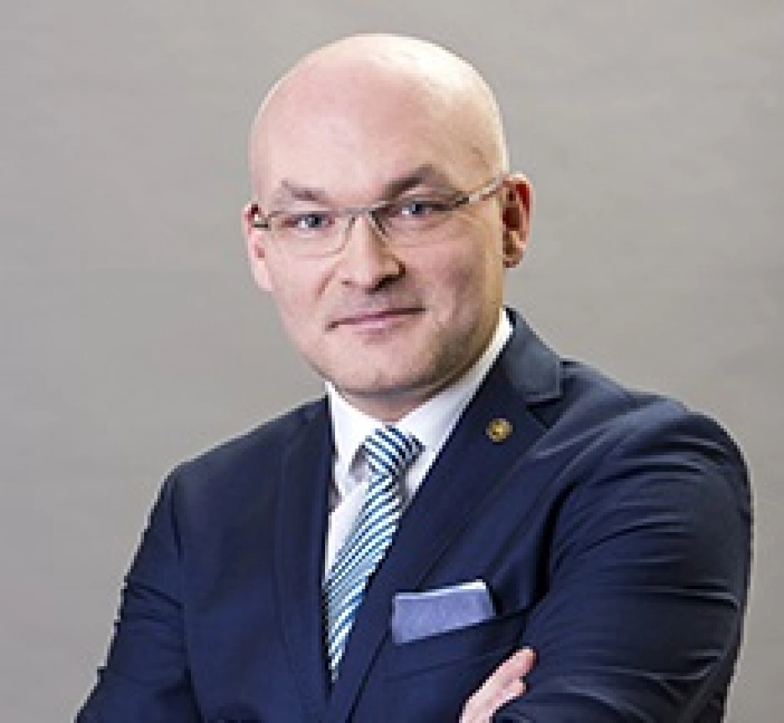 Mężczyzna w okularach, w garniturze, z założonymi rękami, uśmiecha się do kamery.