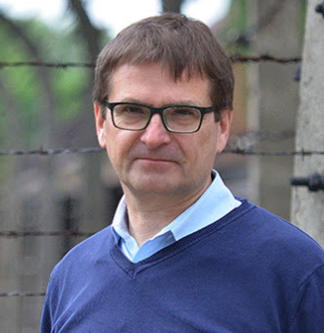 Mężczyzna w okularach stoi przy ogrodzeniu z drutu kolczastego.