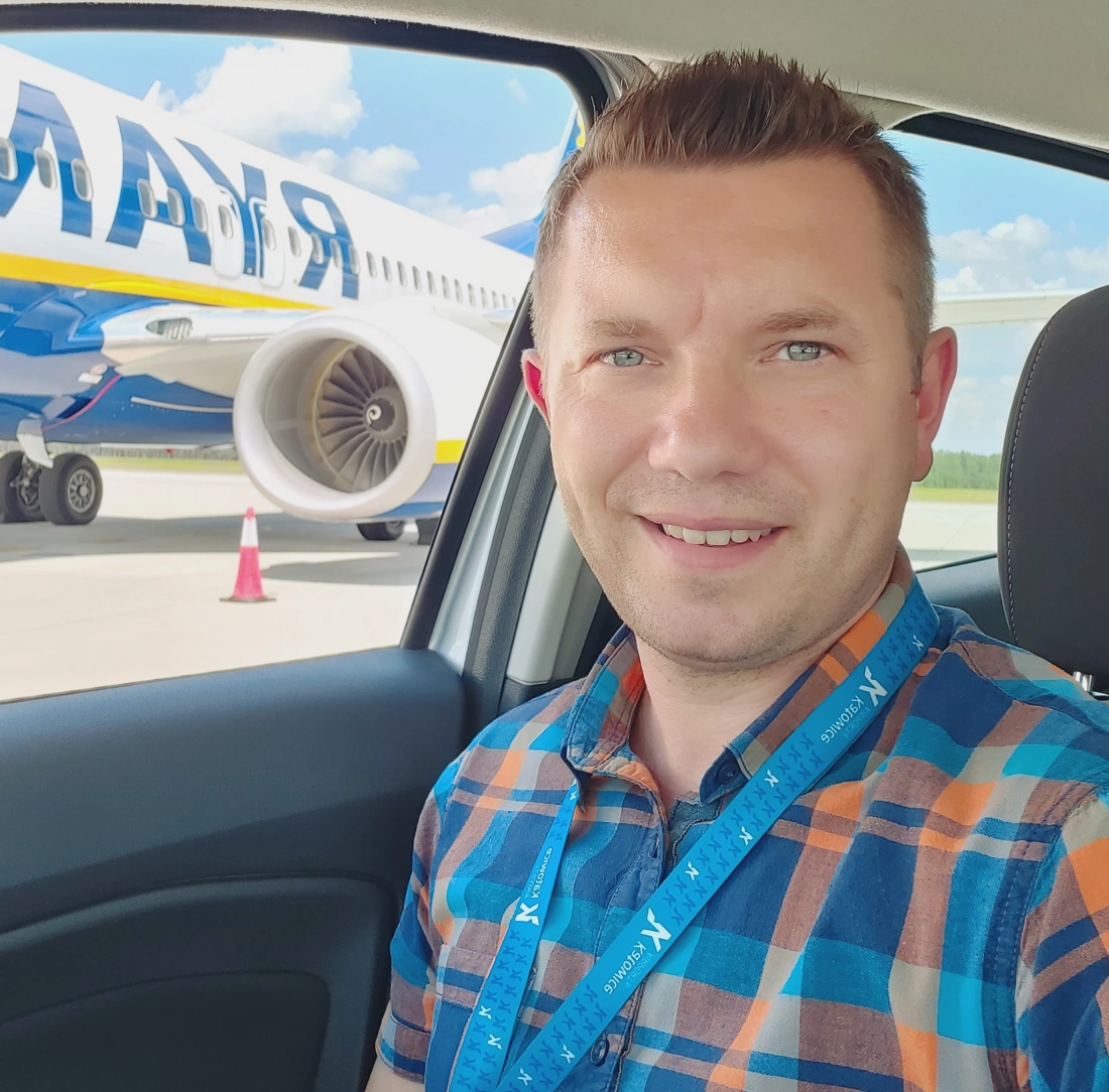 Mężczyzna w samochodzie z uśmiechem, w tle widoczny samolot linii lotniczej.