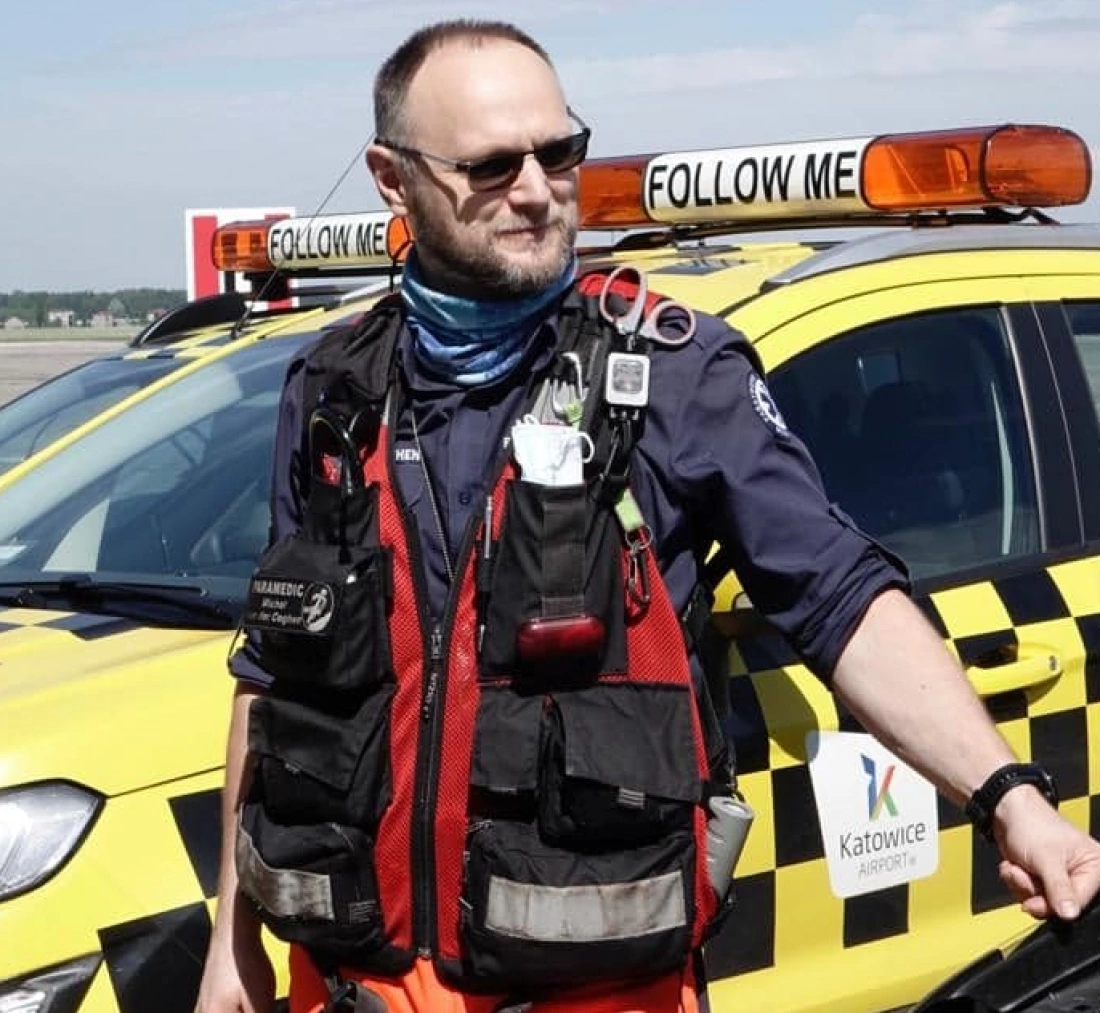 Mężczyzna w specjalistycznym ubiorze ratownika stoi obok żółto-czarnego pojazdu, z napisem "FOLLOW ME" na dachu.