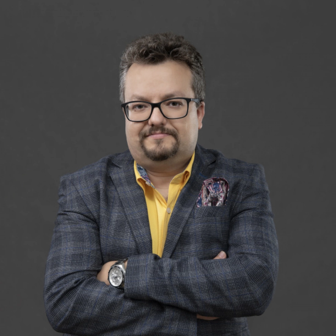 Mężczyzna z okularami i zarostem, ubrany w szary marynarkę i ż&oacute;łtą koszulę, ze złączonymi rękami na piersi.