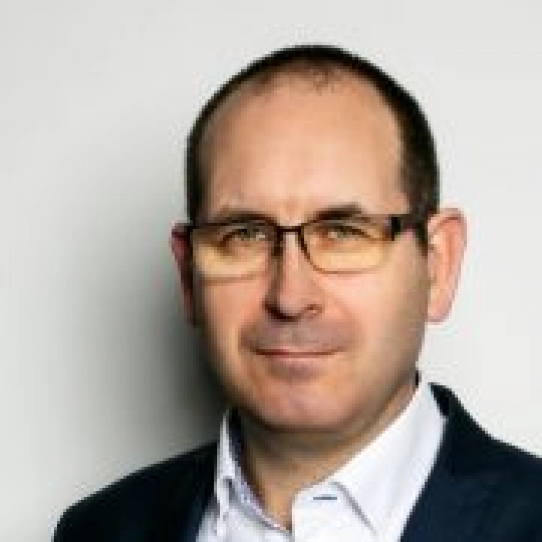 Mężczyzna w okularach, w białej koszuli i ciemnej marynarce, z poważnym wyrazem twarzy.