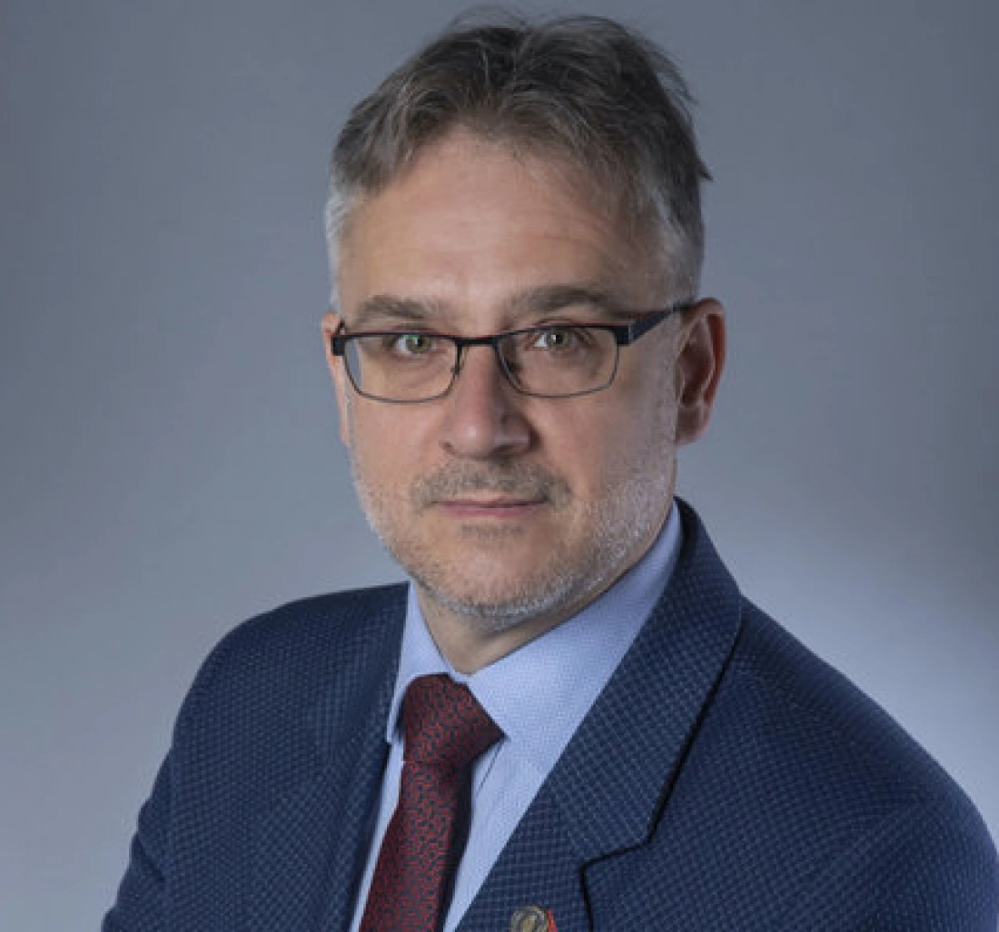 Mężczyzna w okularach, ubrany w granatowy garnitur, pozujący na neutralnym tle.