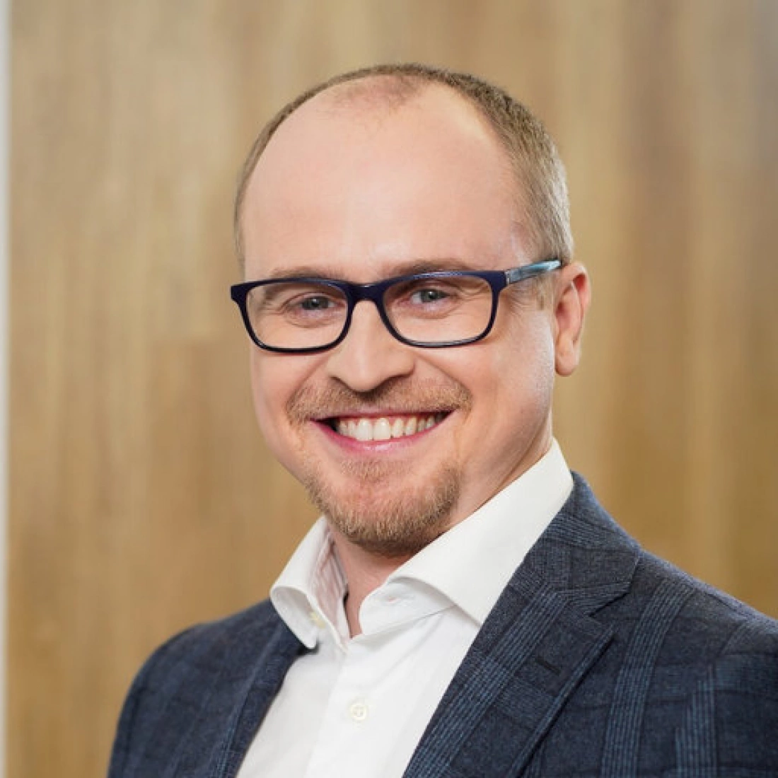 Mężczyzna uśmiechający się w okularach, w eleganckim stroju, stojący na tle drewnianej ściany.