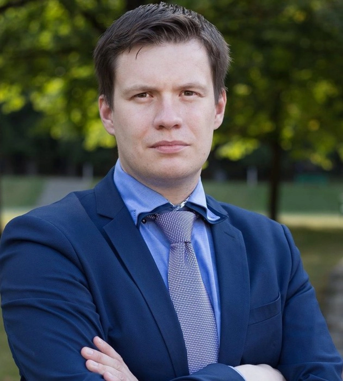 Mężczyzna w garniturze z założonymi rękami, na tle zielonej scenerii.