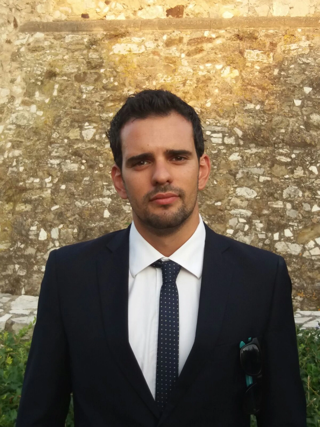 Mężczyzna w eleganckim garniturze, stojący przed kamiennym tłem.