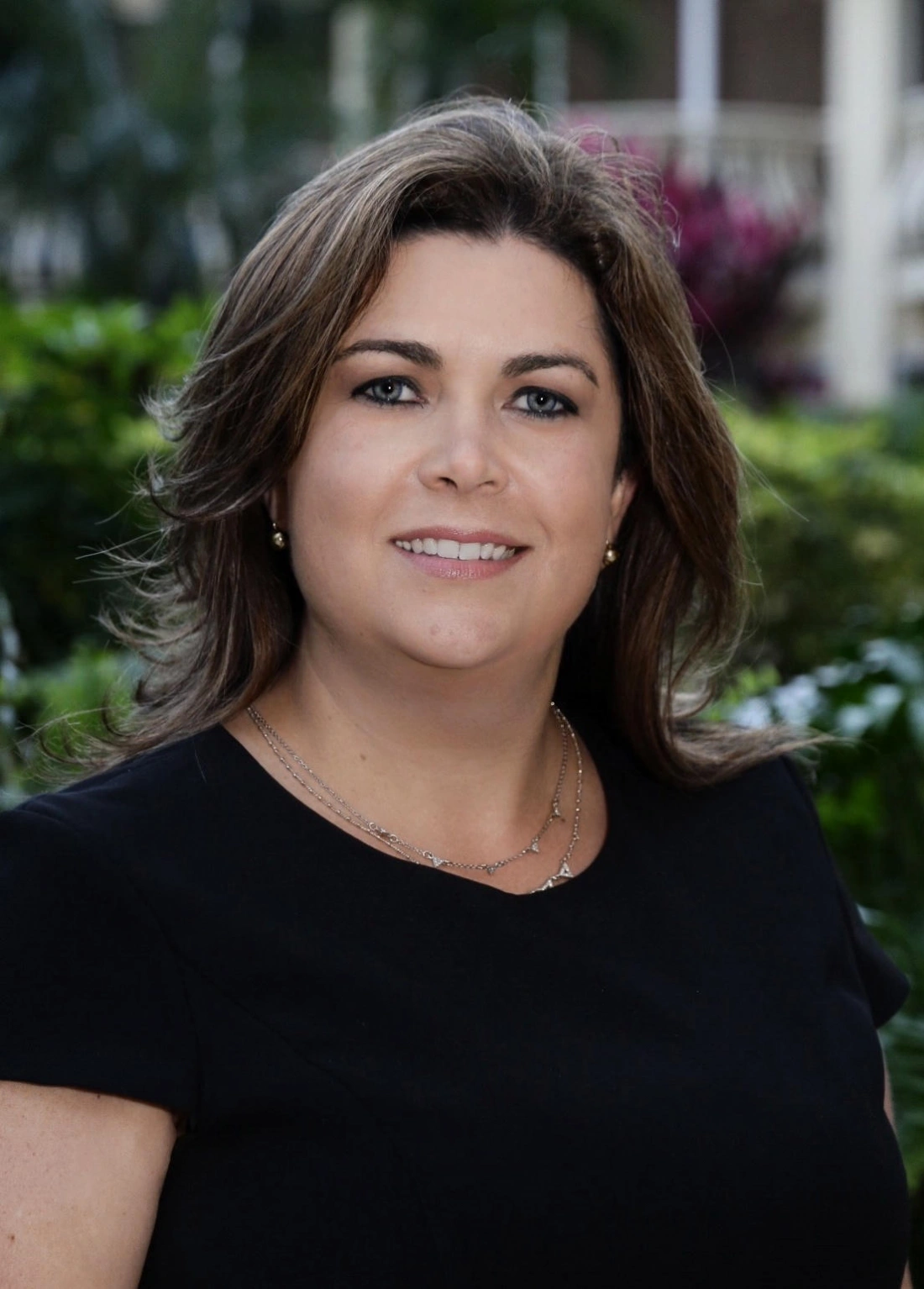 Kobieta w czarnej sukience, uśmiechająca się na tle zieleni.