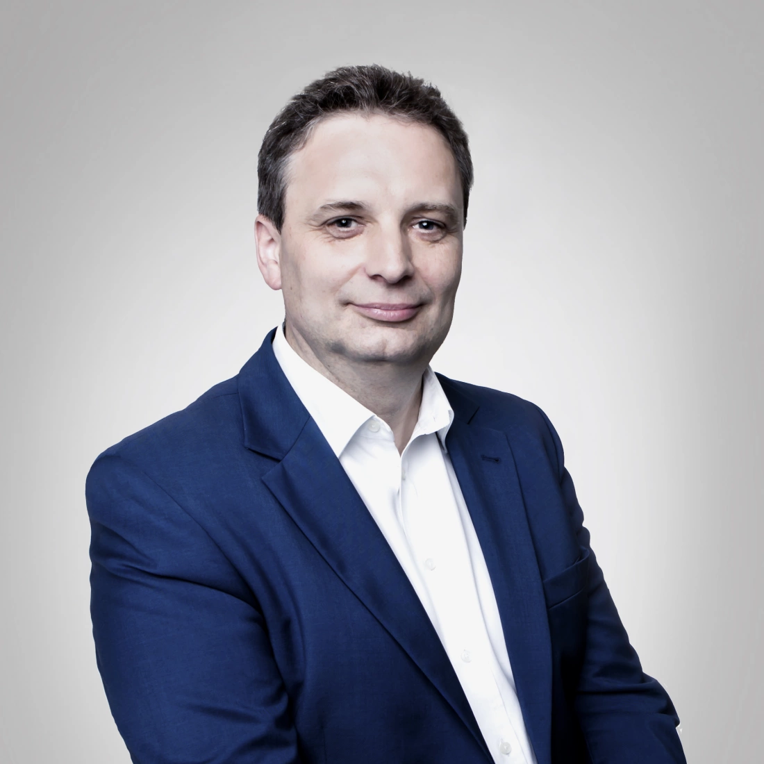 Mężczyzna w garniturze z lekkim uśmiechem, na neutralnym tle.