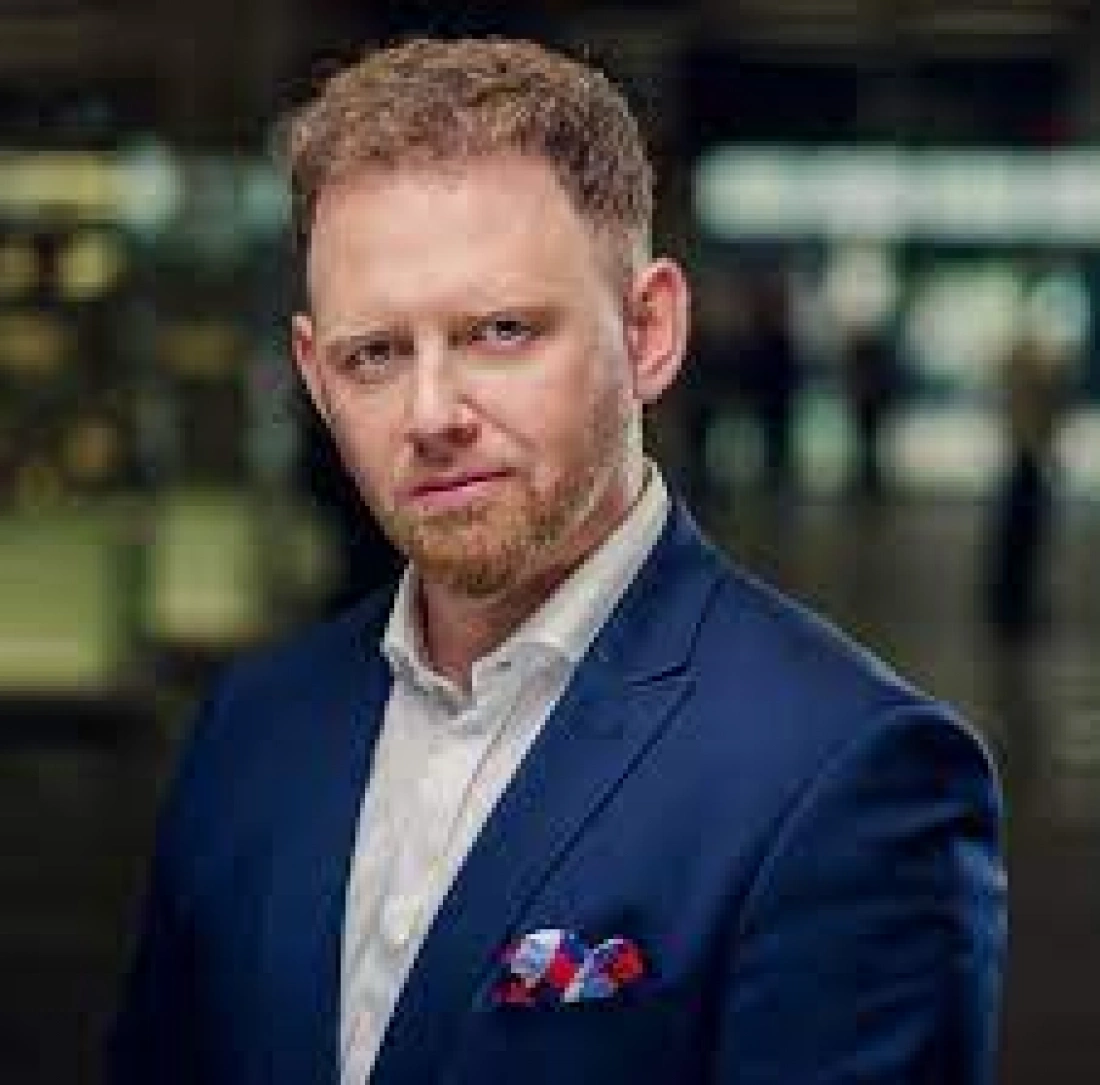 Mężczyzna w granatowym garniturze z okularami i czerwono-niebieską poszetką.