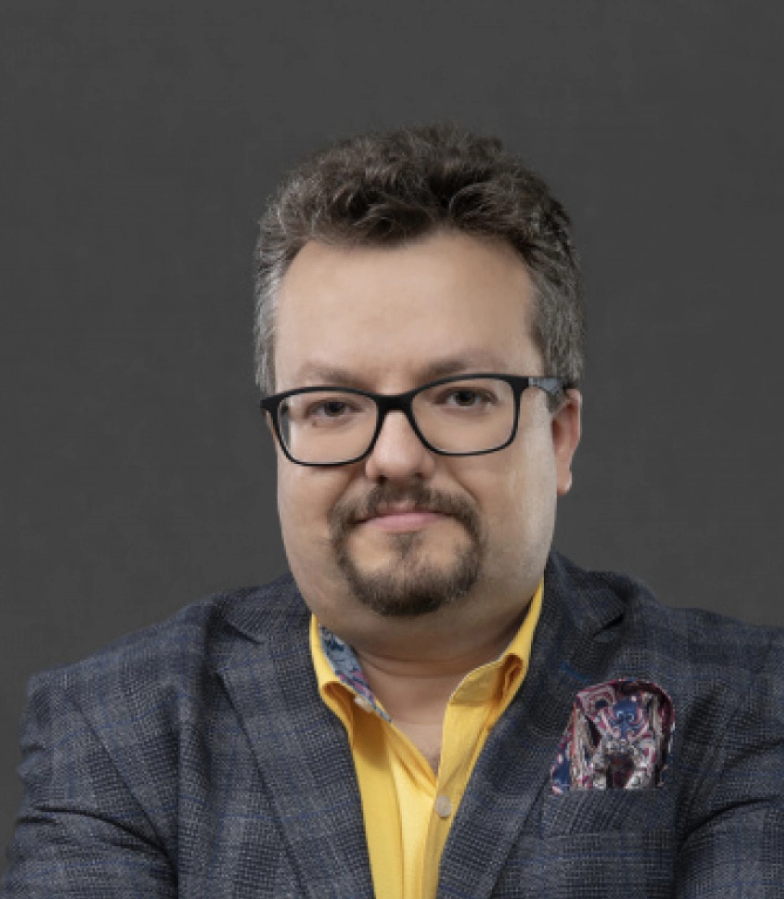 Mężczyzna w okularach, w szarej marynarce i ż&oacute;łtej koszuli, uśmiecha się na tle ciemnego tła.