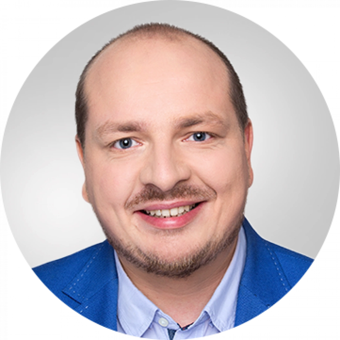 Mężczyzna w niebieskim garniturze z uśmiechem, na tle szarej ściany.