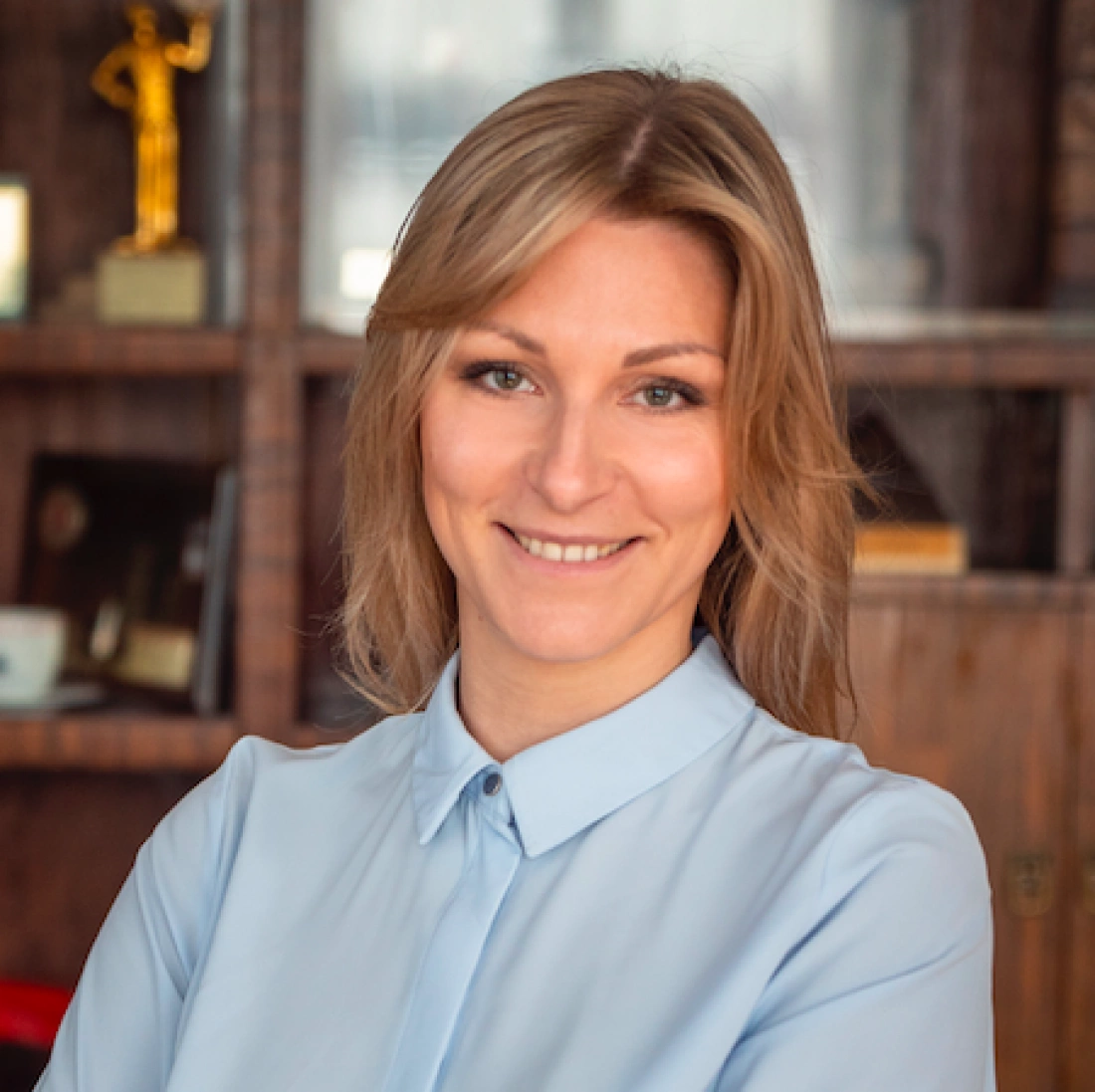 Kobieta w jasnej koszuli uśmiecha się w biurze, w tle półka z trofeami.