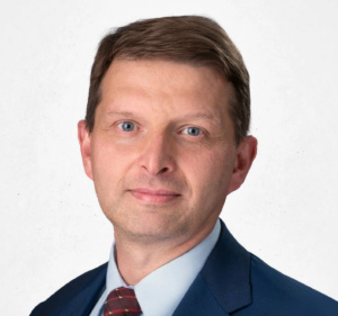 Mężczyzna w garniturze z krótkimi, brązowymi włosami i niebieskimi oczami, uśmiechający się na tle neutralnym.