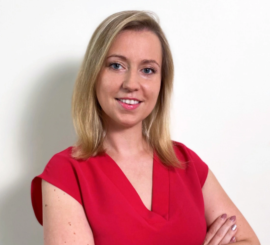 Kobieta z blond włosami w czerwonej bluzce, uśmiechnięta, z założonymi rękami. W tle jasna ściana.