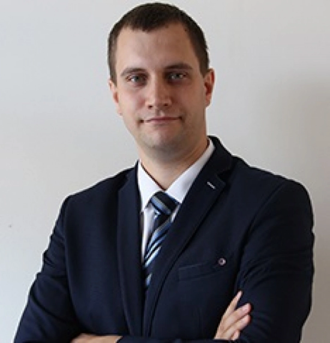 Mężczyzna w garniturze z założonymi rękami na tle jasnej ściany.