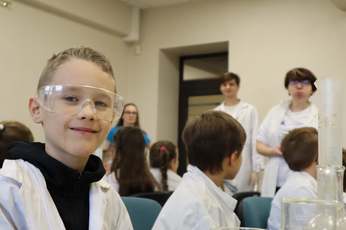 Chłopiec w okularkach ochronnych uśmiecha się, siedząc w sali pełnej dzieci w laborkach chemicznych. W tle dorośli prowadzą zajęcia.