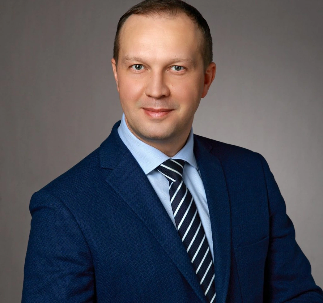 Mężczyzna w garniturze z krawatem, uśmiechający się do kamery na tle o stonowanej fakturze.