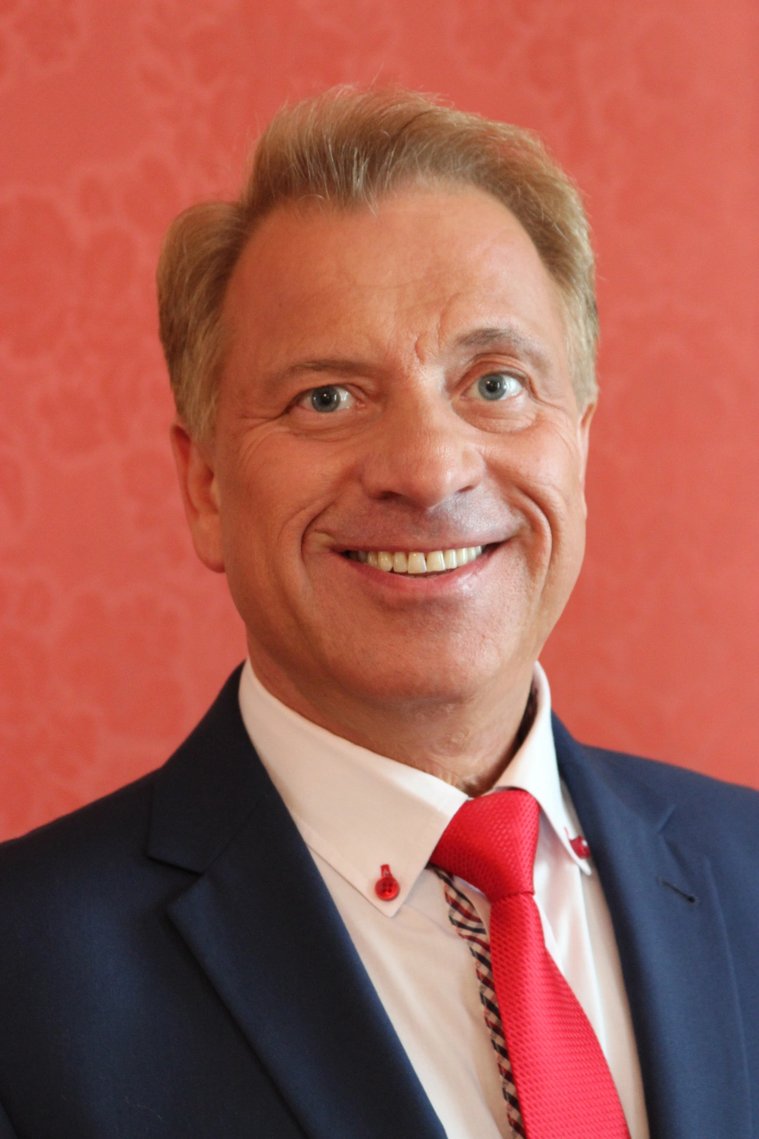 Mężczyzna uśmiechający się w eleganckim garniturze i czerwonym krawacie na tle r&oacute;żowej ściany.