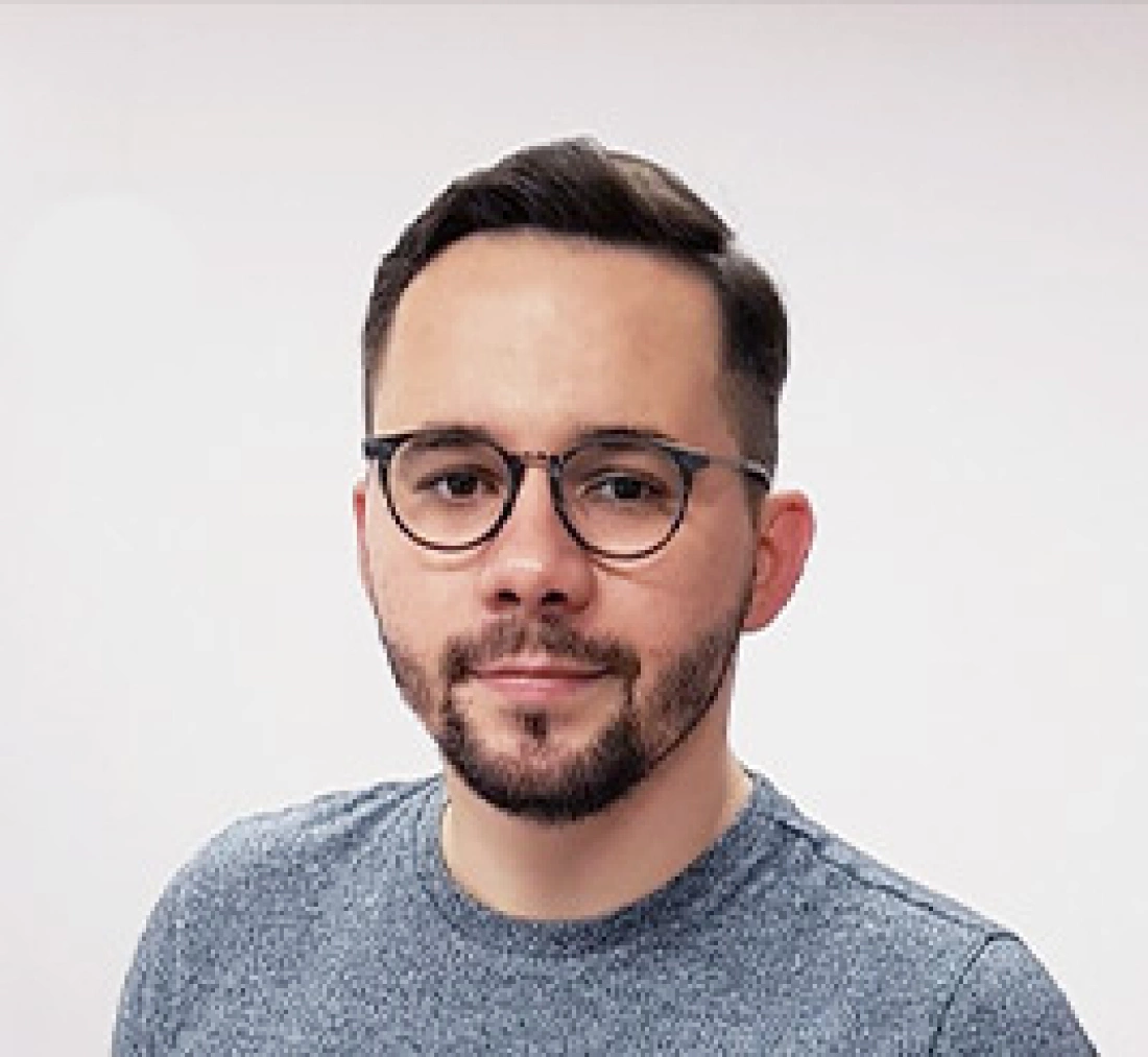 Mężczyzna w okularach, ubrany w szary t-shirt, stoi na neutralnym tle.