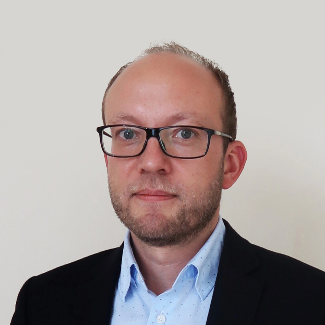 Mężczyzna w okularach, ubrany w garnitur, stoi na jasnym tle.