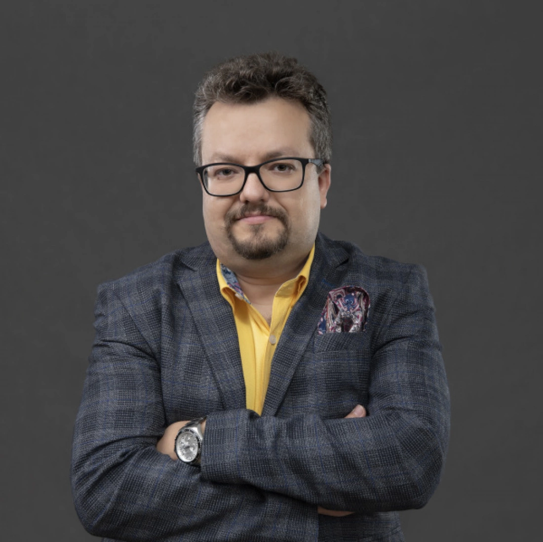 Mężczyzna z okularami w marynarce stoi z założonymi rękami na szarym tle.