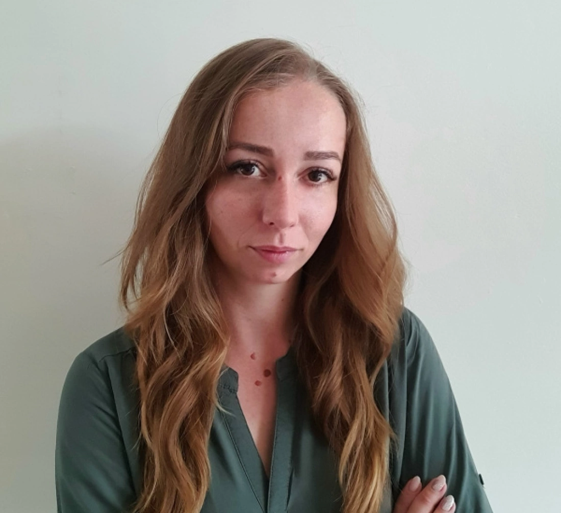 Kobieta z długimi włosami w zielonej bluzce, z poważnym wyrazem twarzy, stojąca na tle jasnej ściany.