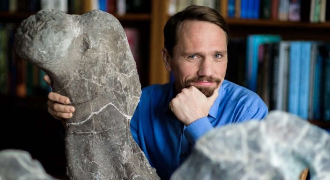 Mężczyzna w niebieskiej koszuli stoi obok dużego odłamu skały, otoczony książkami.