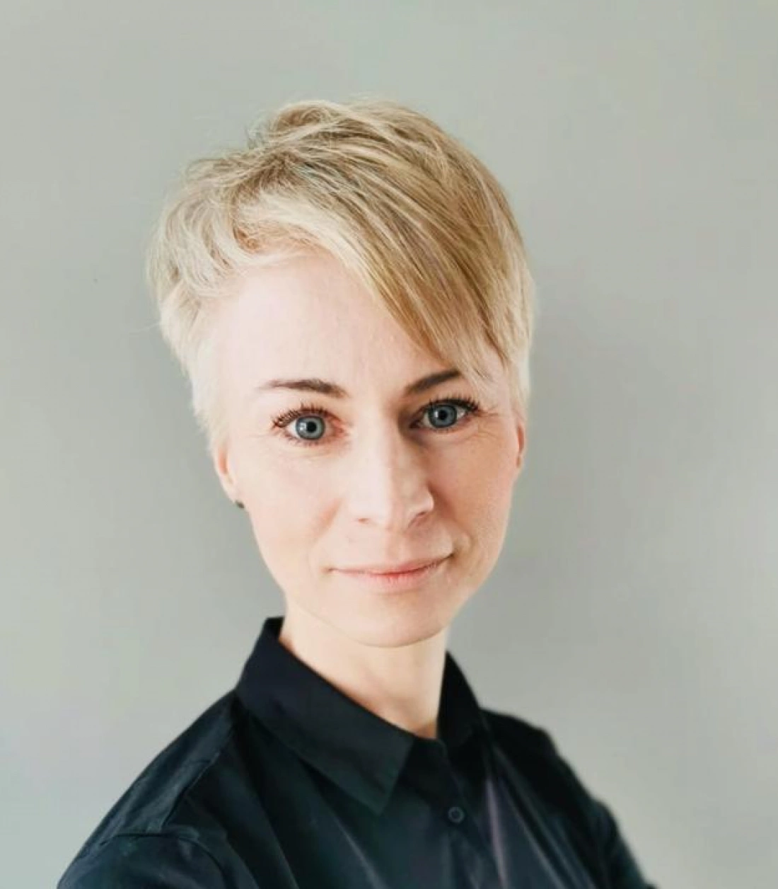 Kobieta z krótkimi blond włosami, w czarnej koszuli, patrzy prosto w obiektyw na tle szarej ściany.