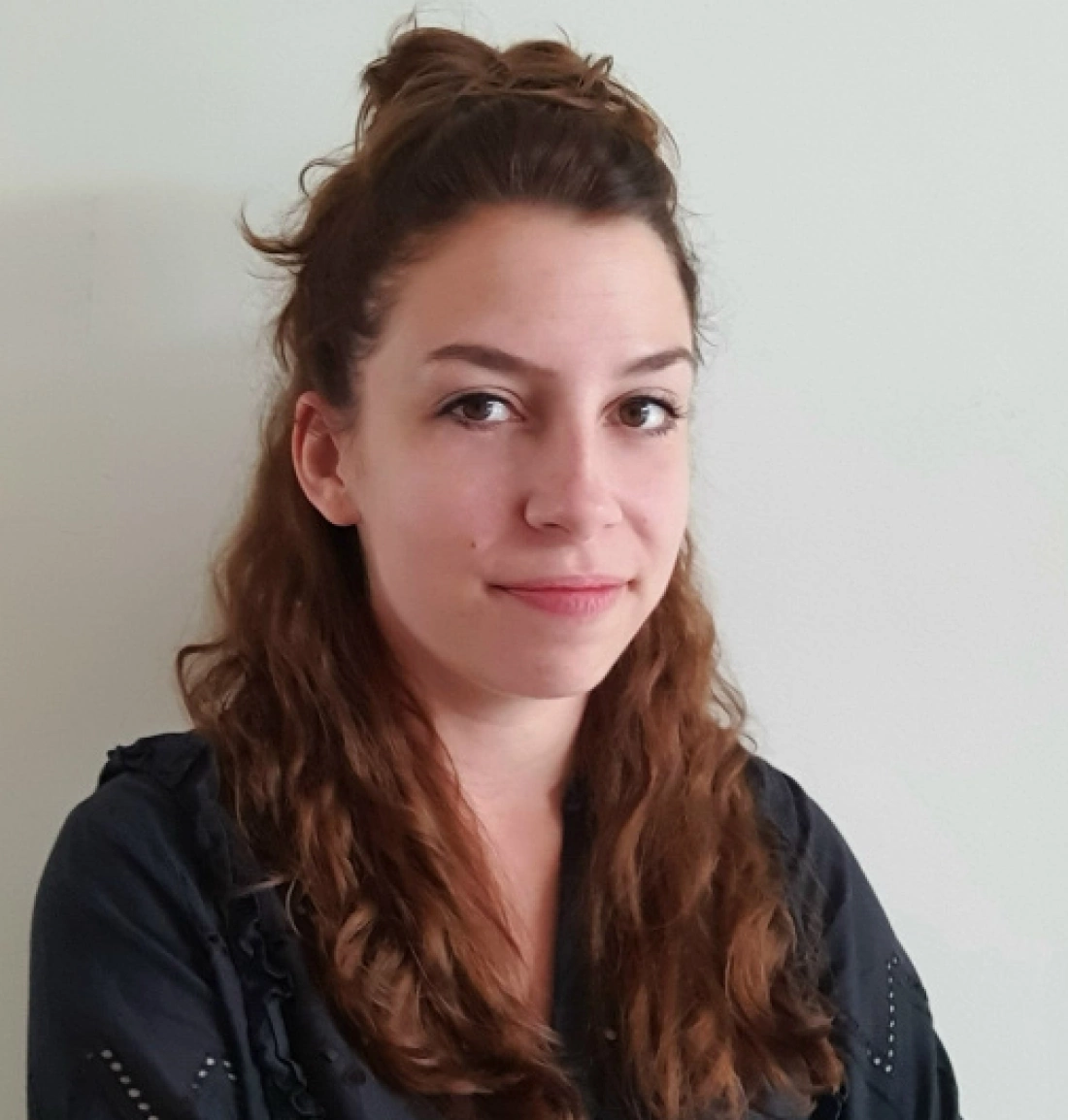 Młoda kobieta z długimi, kręconymi włosami, ubrana w ciemną bluzę, stojąca na jasnym tle.