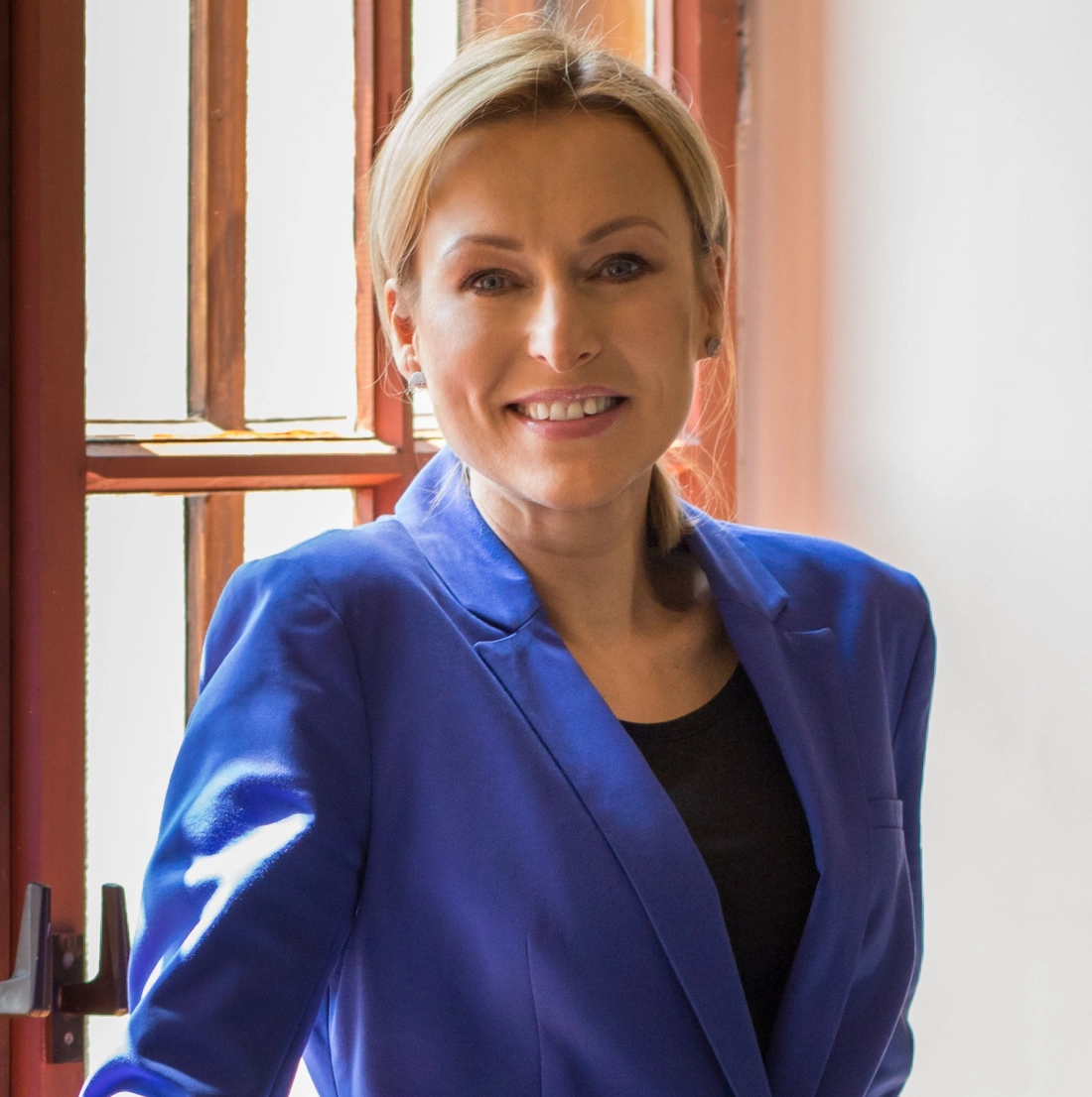 Kobieta w błękitnej marynarce stoi przy oknie, uśmiechając się.
