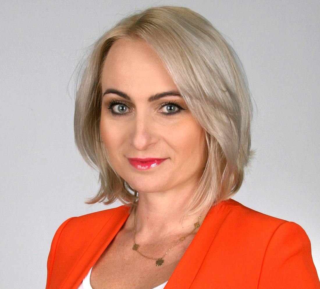 Portret kobiety z jasnymi włosami, ubraną w pomarańczową marynarkę, na tle szarym.